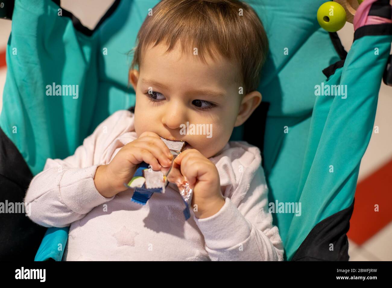little cute hungry toddler girl sitting in a stroller and eating a lozenge candy bar. close-up, soft focus. Stock Photo