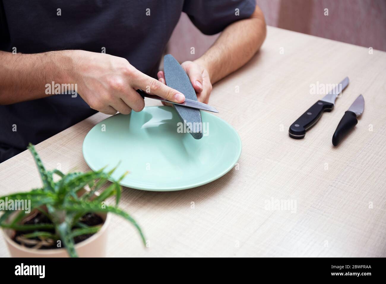 Close-up strong male hands sharpen a kitchen metal knife with a grindstone. Home household knife sharpening. Stock Photo