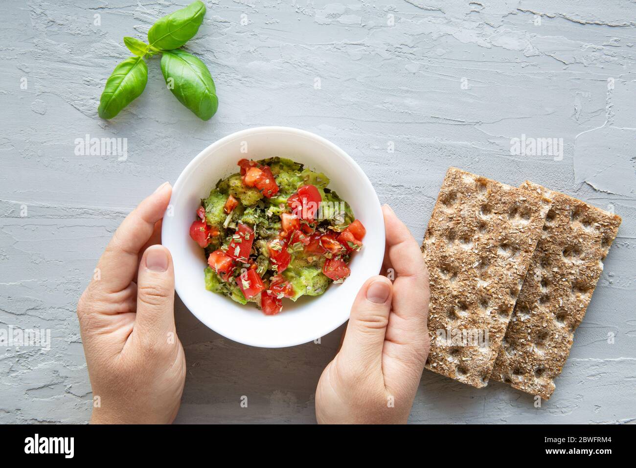 Female hands hold tasty guacamole bowl with salsa on grey concrete background, top view. Stock Photo
