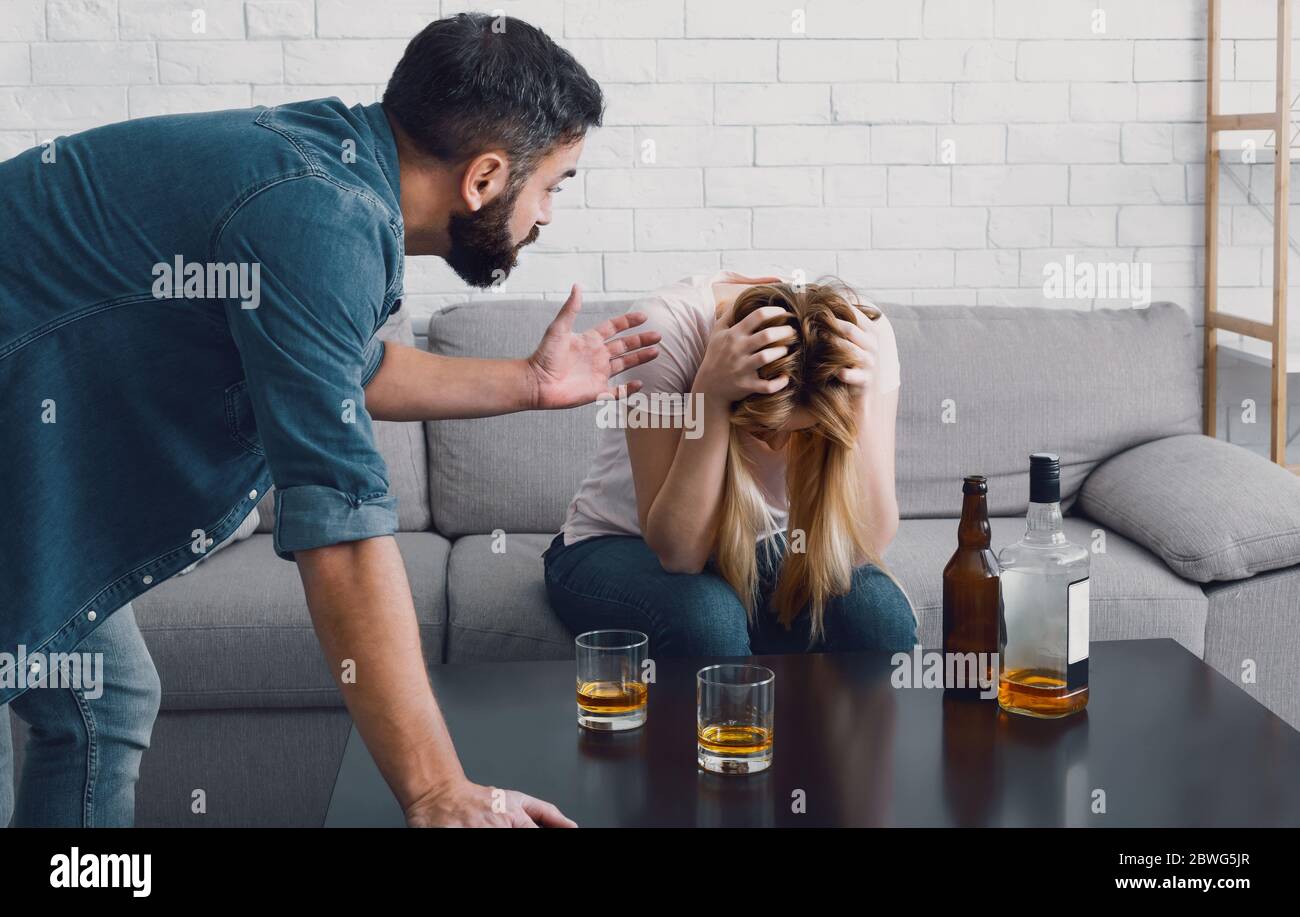 Drunken scandal. Woman holds her head and sitting on sofa, angry man swears with wife Stock Photo