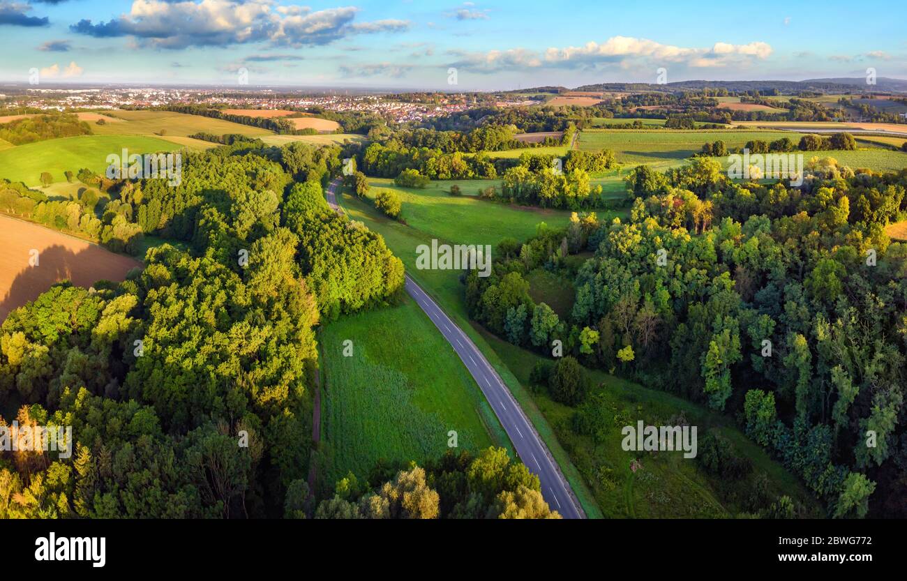 Idyllic German landscape shot from above in early morning sunlight: meadows and forests with blue sky and a road leading to a small town on the horizo Stock Photo
