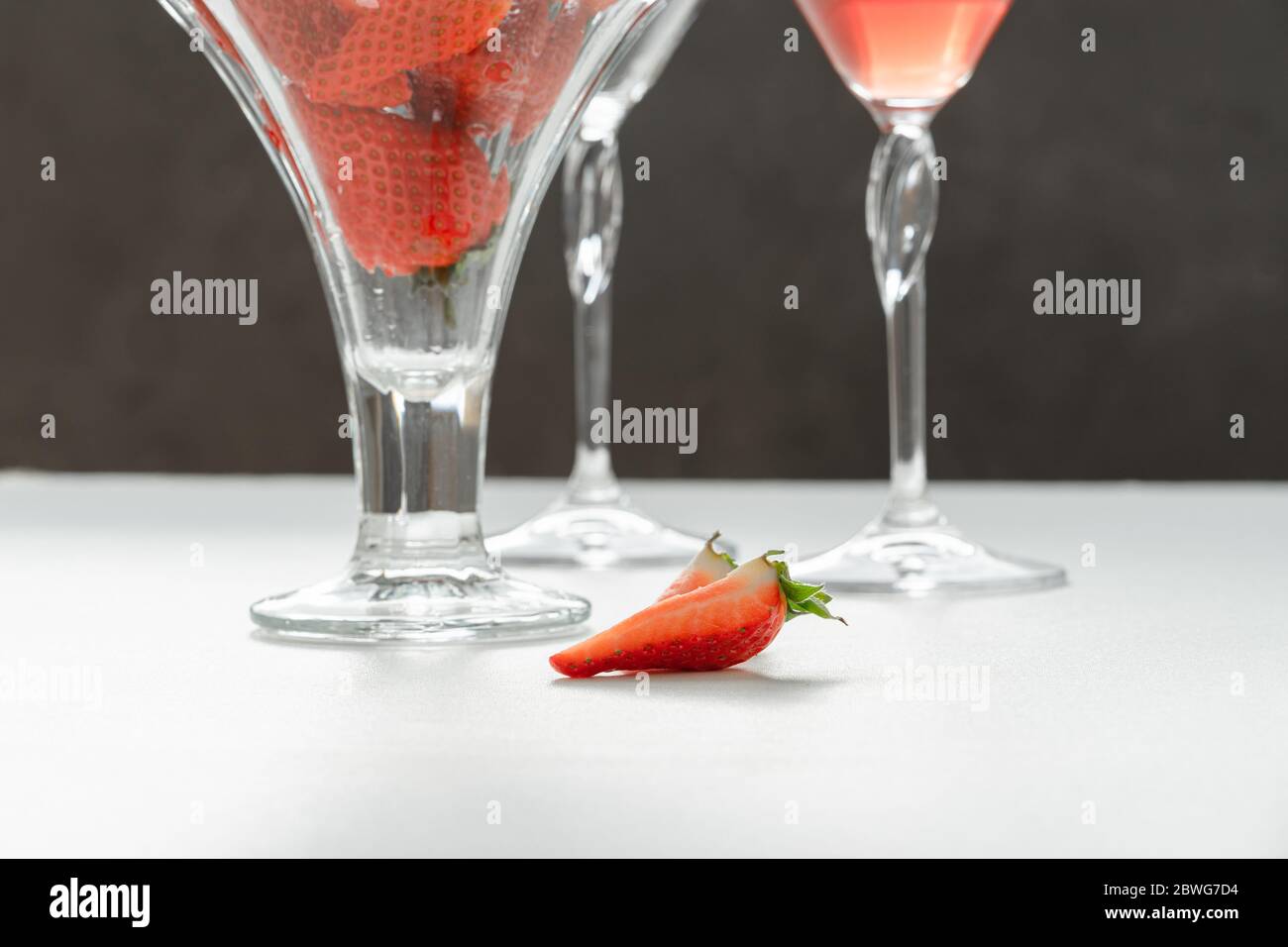Summer arrangement. Pieces of strawberry lie on a white table surface, behind them - glasses with strawberries and a red drink. The concept of holiday Stock Photo