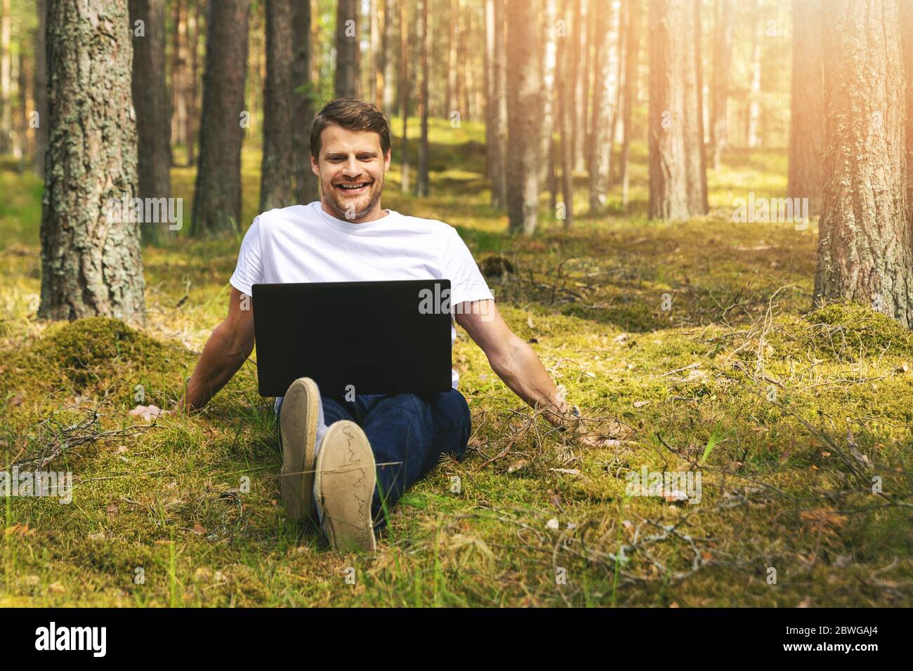 young smiling man sitting in the moss and using laptop in the forest Stock Photo