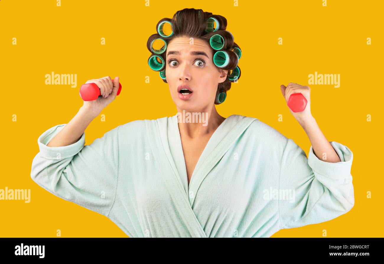 Shocked Housewife Exercising With Dumbbells Training At Home, Yellow Background Stock Photo