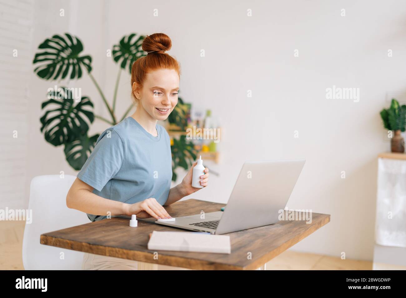 Smiling young redhead woman is wiping laptop computer with sanitizer before starting work  Stock Photo