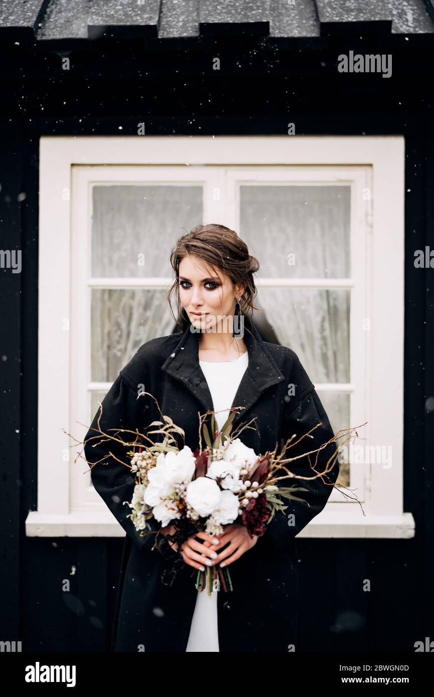 Portrait of a bride in a white silk wedding dress and a black coat with a bride's bouquet in her hands. Black wooden house with a white window Stock Photo