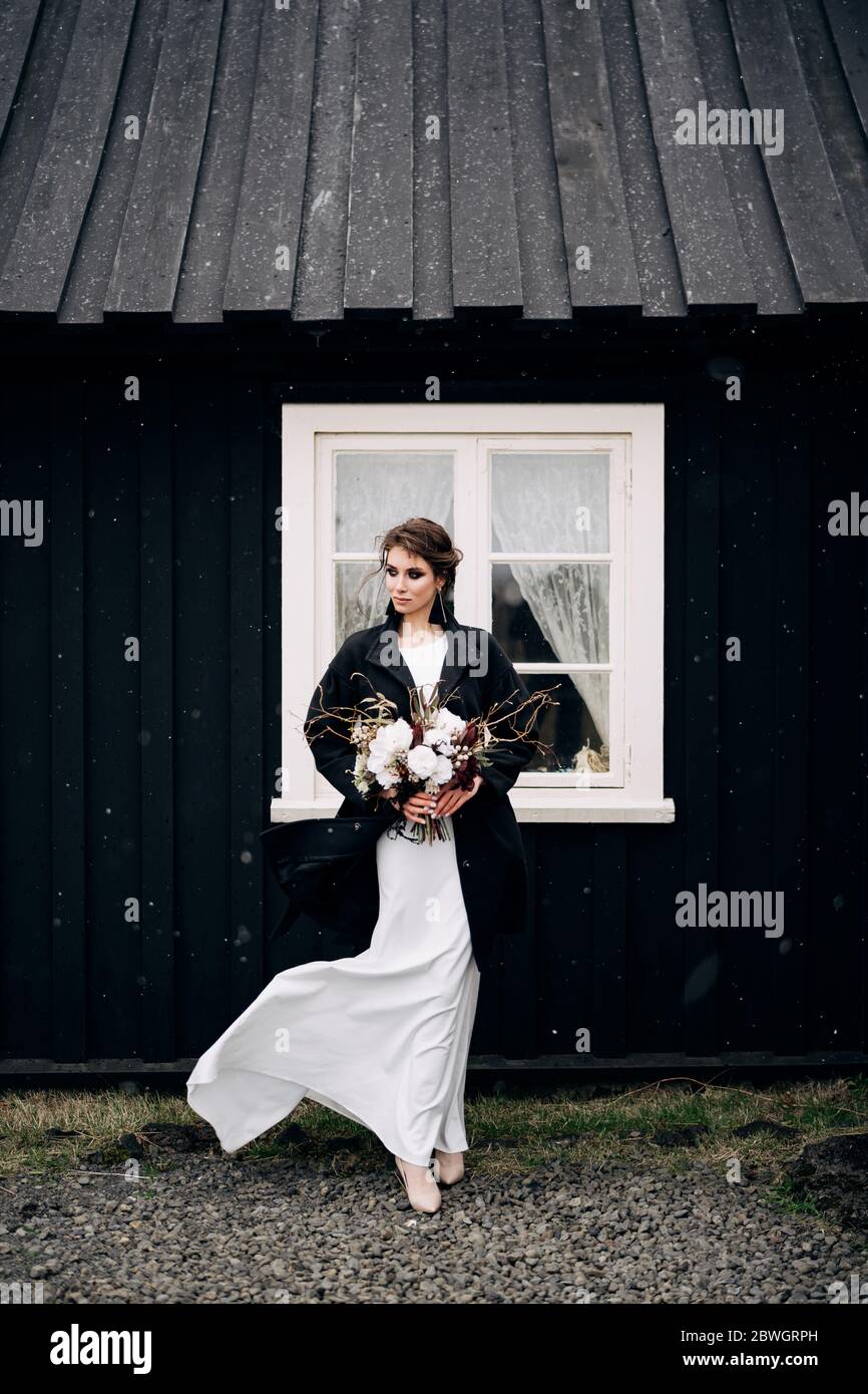 Portrait of a bride in a white silk wedding dress and a black coat with a bride's bouquet in her hands. Black wooden house with a white window Stock Photo