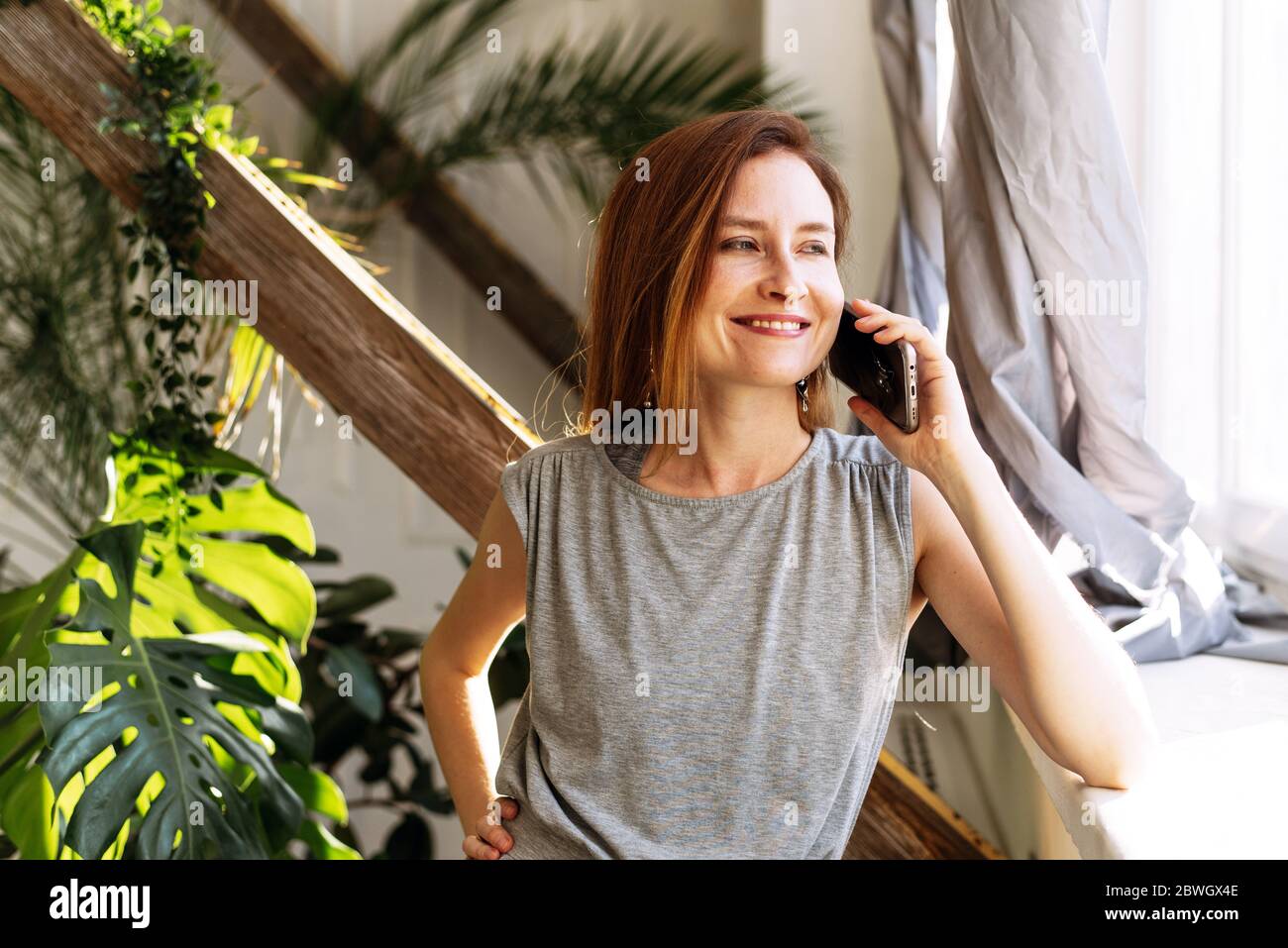 A pretty woman speaks at home on the phone with relatives or friends on a background of diverse plants. Stock Photo