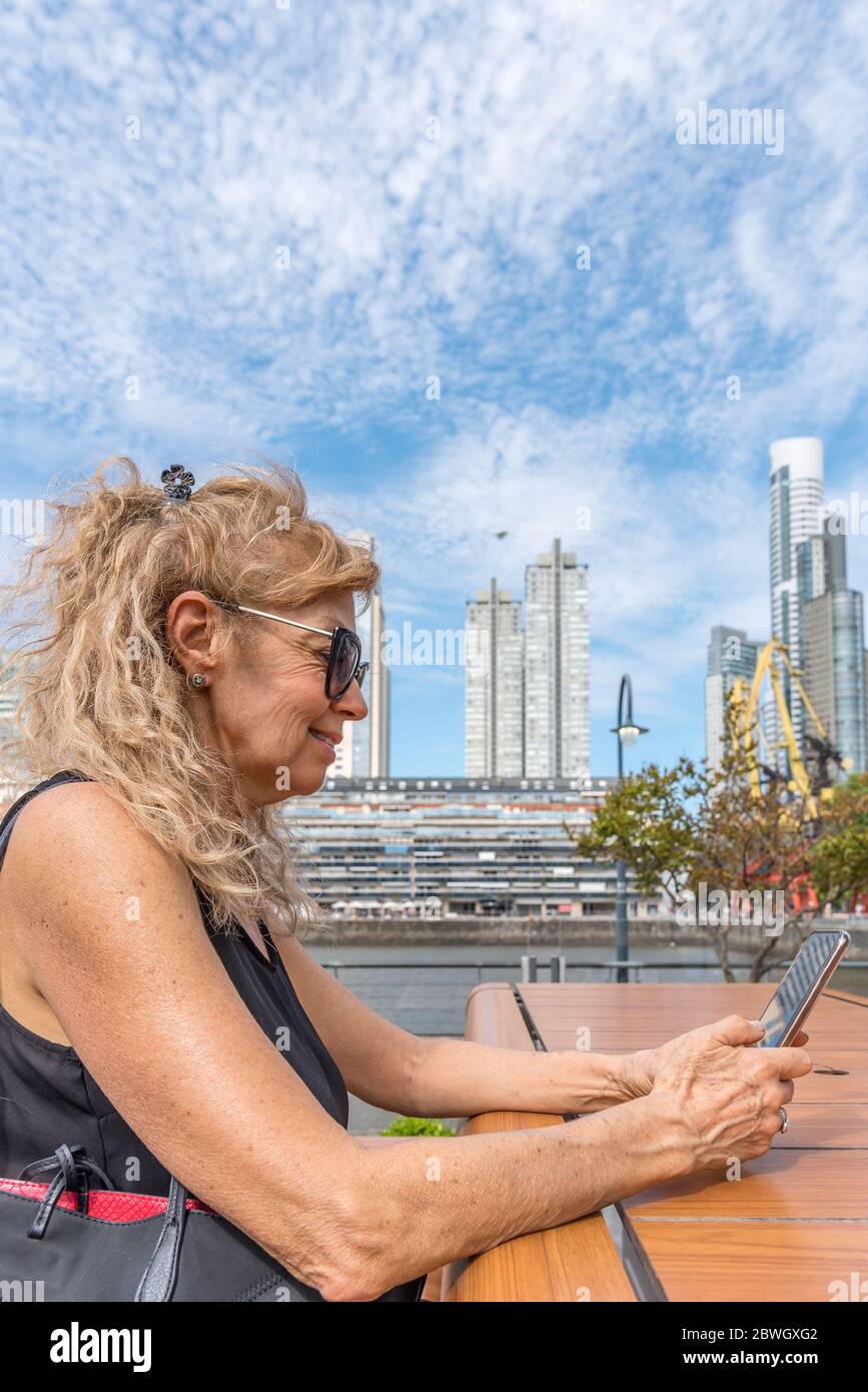 Adult blonde woman smiling as she looks at her mobile phone with the buildings of Puerto Madero, Buenos Aires in the background. Stock Photo