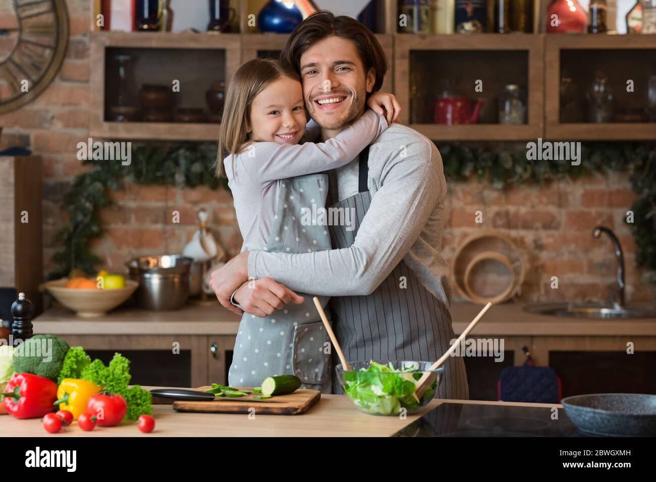 Family Love. Cheerful little girl hugging with her happy dad in kitchen Stock Photo