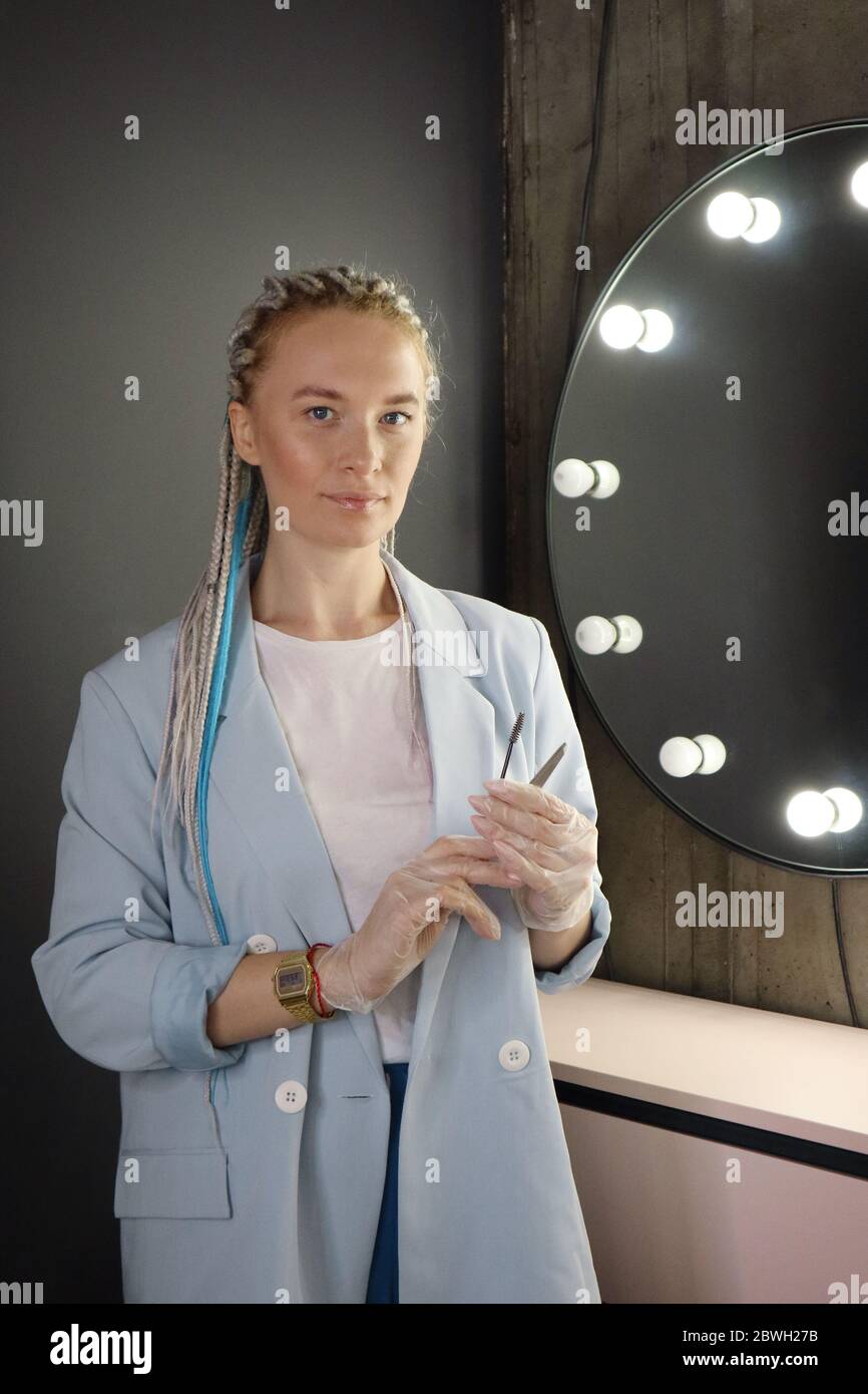 Beautiful young woman brow designer with a blue-and-white dreadlocks,  tweezers and eyebrow brush in hand near the mirror with bulbs Stock Photo