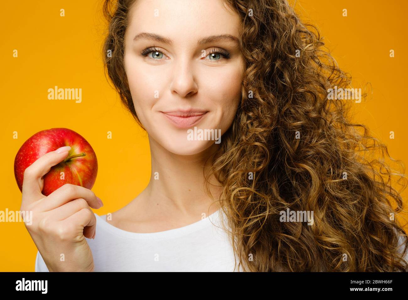 Closeup portrait of pretty caucasian girl with curly hair and red apple isolated on colorful yellow background Stock Photo