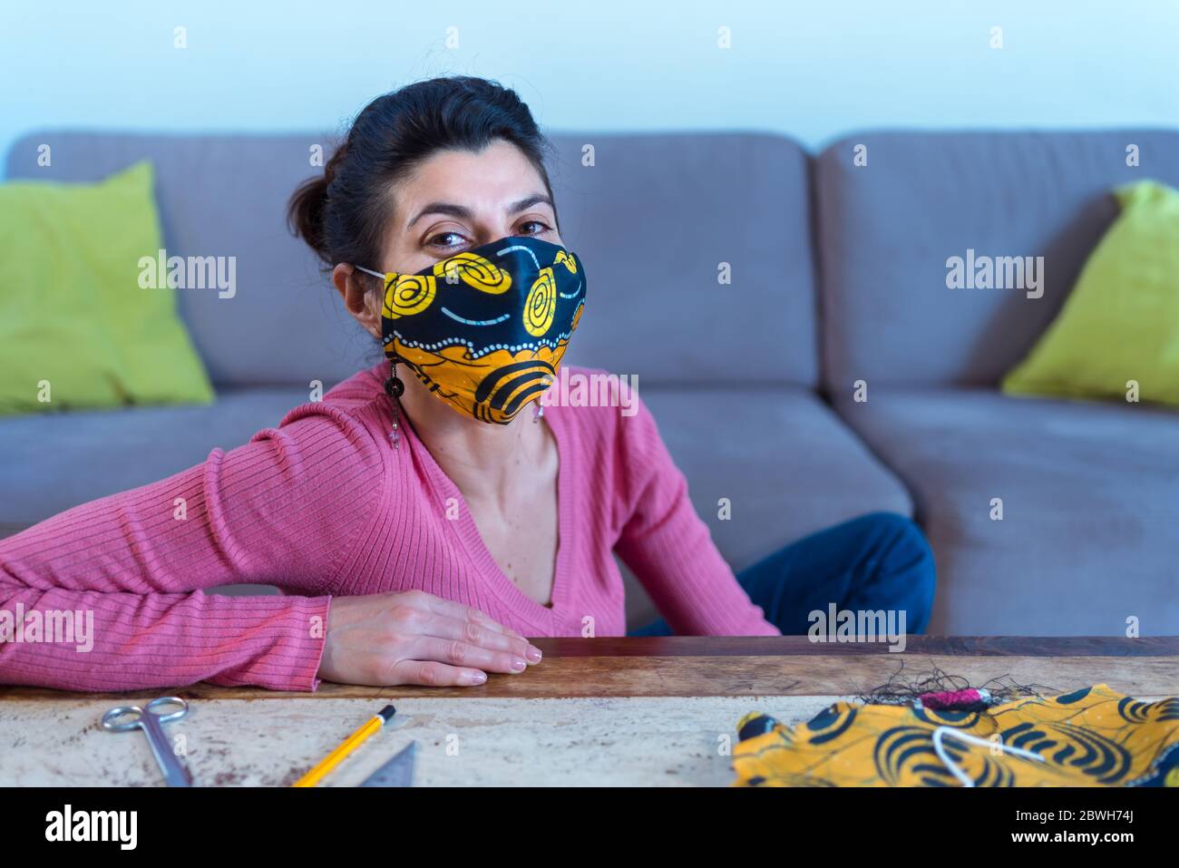 A happy young girl sitting by the sofa wears a cloth face mask she has hand made with a sewing machine. Stock Photo