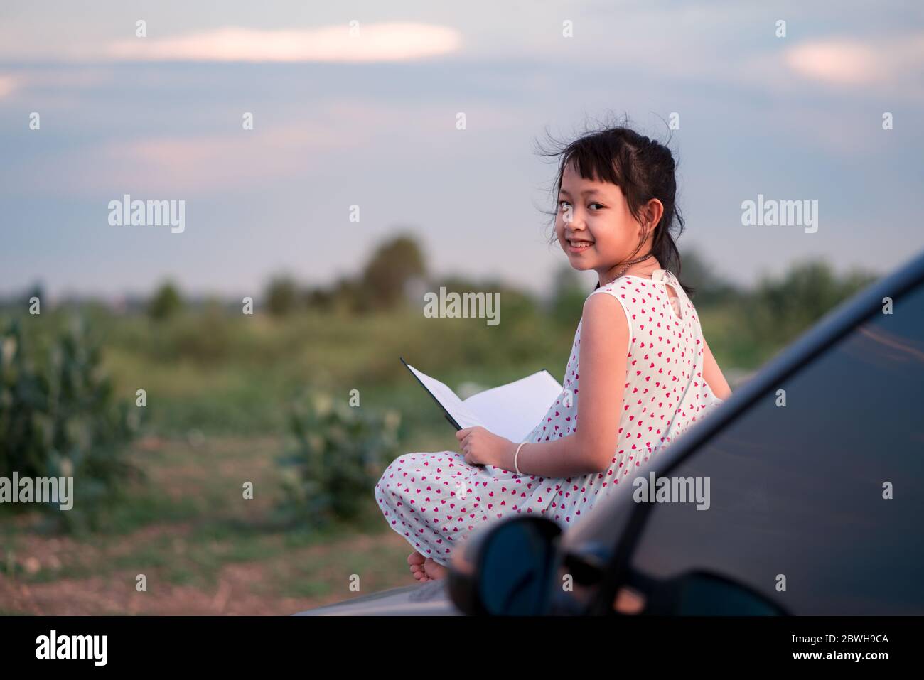 Little child girl  sitting and relaxing on a car reading a book in a green nature background Stock Photo