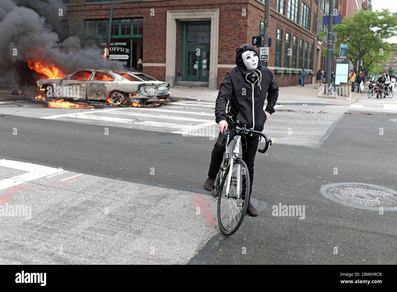 A protester with a Guy Fawkes mask rests on his bike in front of burning police cars in downtown Cleveland, Ohio during the George Floyd protest. Stock Photo