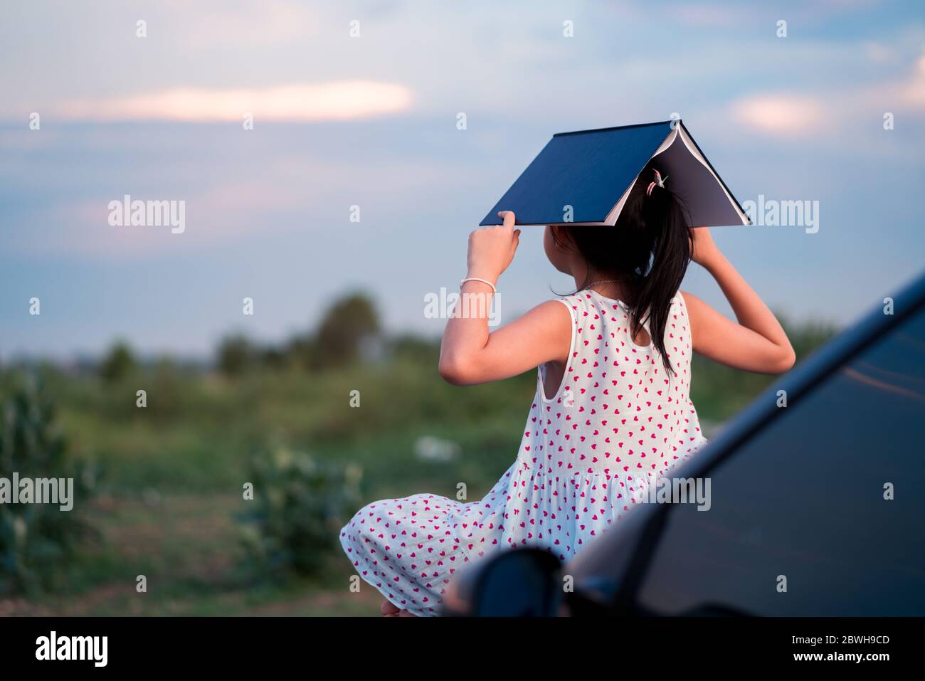 Little child girl  sitting and relaxing on a car reading a book in a green nature background Stock Photo