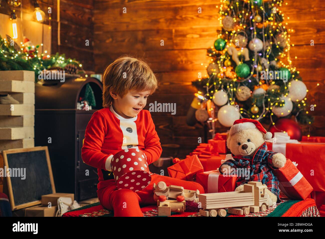 Happy little kid is playing with Christmas gift boxes on Christmas tree background. Kid is waiting for the New Year. Holidays. Stock Photo