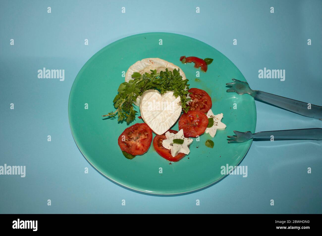 Brie cheese cut in the shape of a heart accompanied by basil leaves and flowers, slices of tomato fruits and slices of star-shaped panela cheese Stock Photo