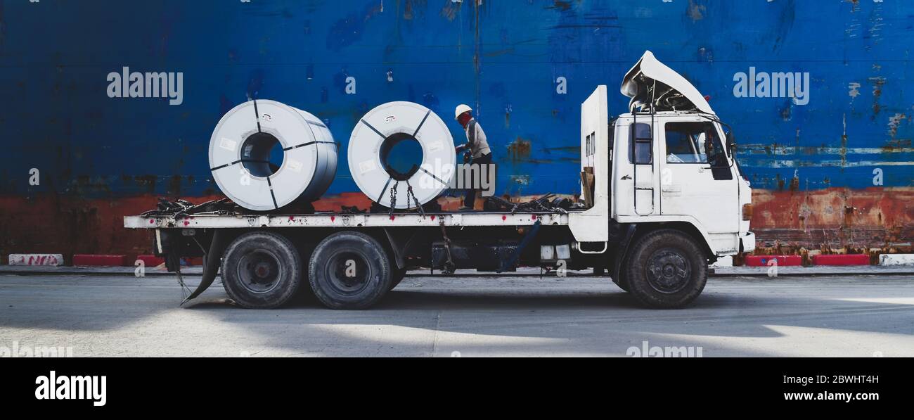 Truck load steel coils alongside big ship. Steel cargo discharged from vessel at seaport. Material logistics concept. Stock Photo