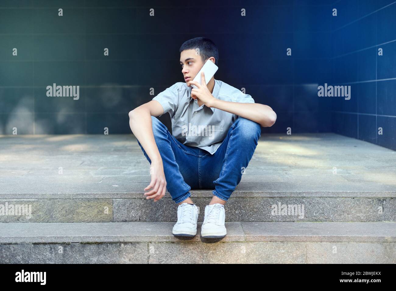 Young teenage boy chatting on his mobile phone outdoors sitting on city steps as he looks away to the side listening to the conversation with a though Stock Photo