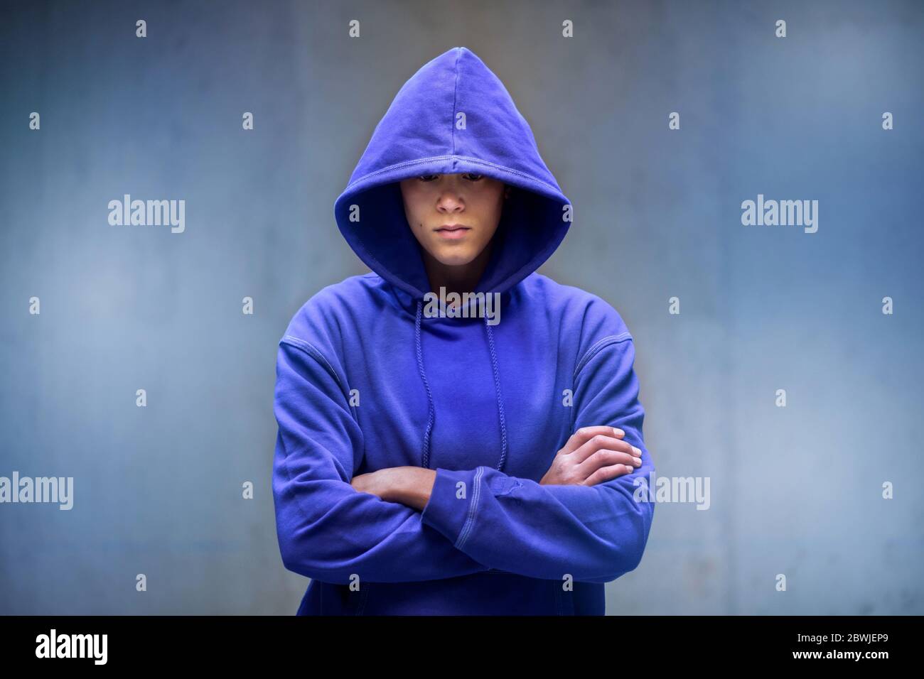 Young man in blue top with hood pulled low over his eyes standing with folded arms facing the camera against a grey wall with copy space Stock Photo