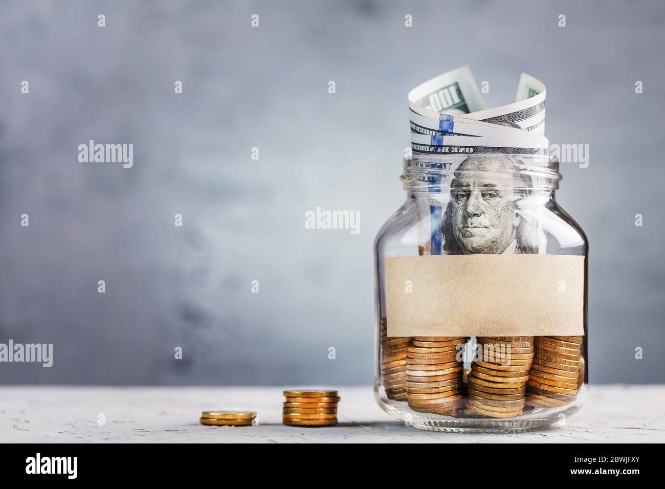 Glass Jar with gold coins, with a hundred-dollar bill and a sticker for text on a gray background with free space for text. Stock Photo