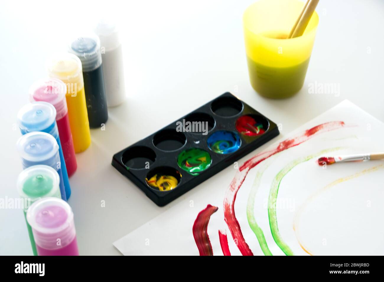 White paper with a child's drawing made of colored paint with several containers of vegetable paint and a plastic cup with water and brushes inside Stock Photo