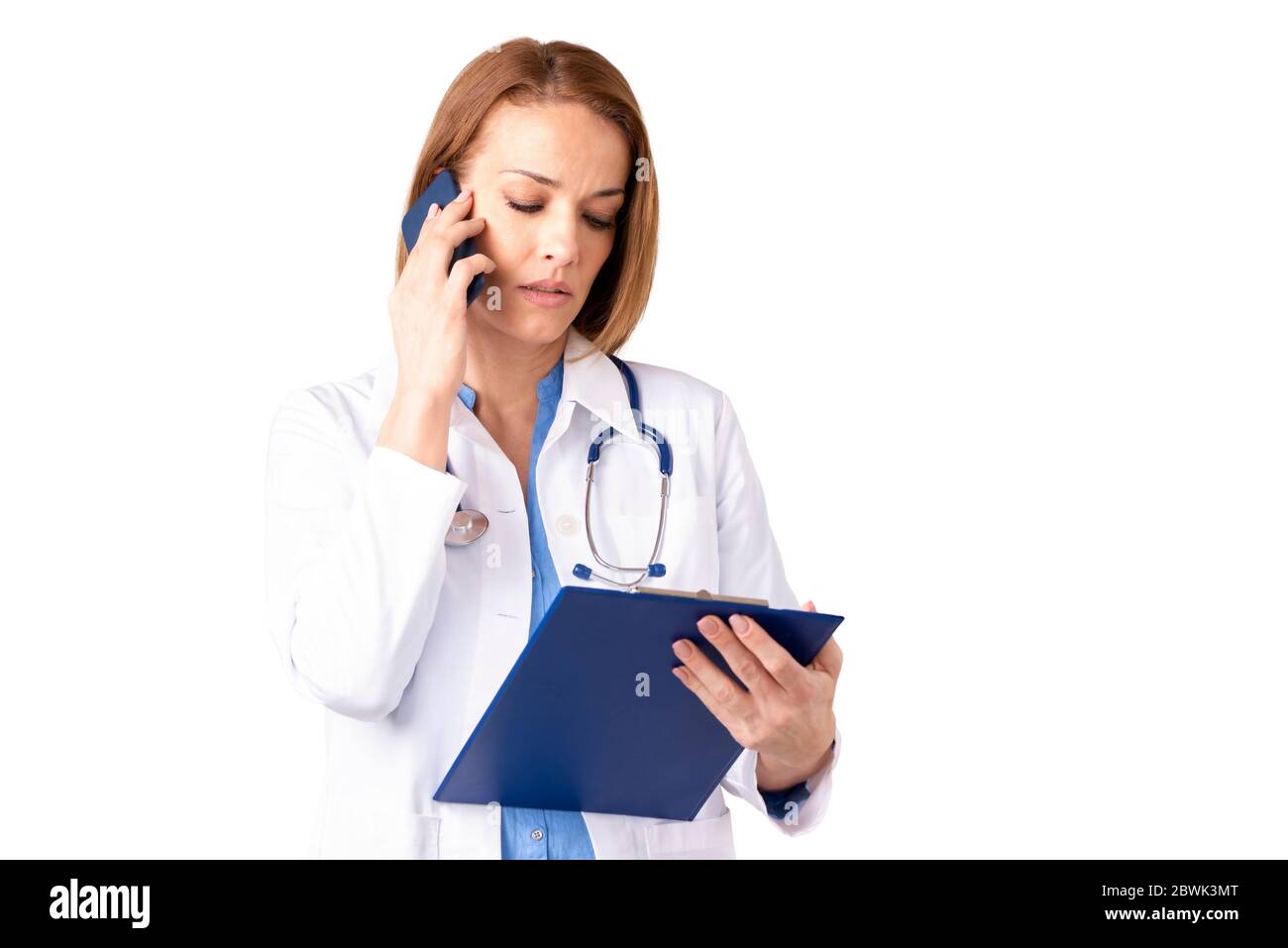 Portrait shot of confident female doctor talking on mobile phone and holding clipboard in her hand while standing at isolated white background. Stock Photo