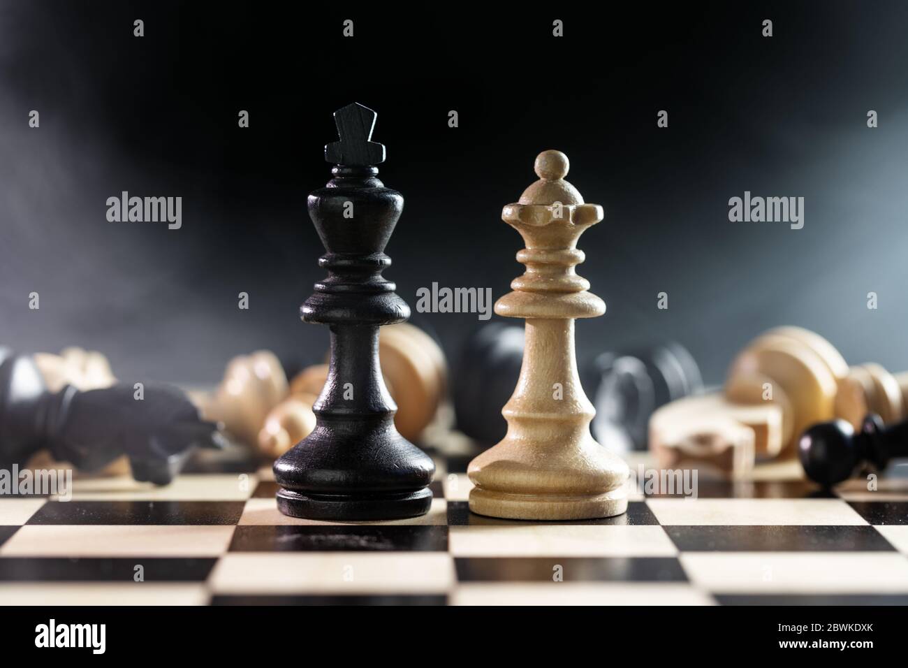 Chessmen queen and king in front of the battlefield of fallen chess pieces sacrifices, black background with smoke and copy space, selected focus, nar Stock Photo