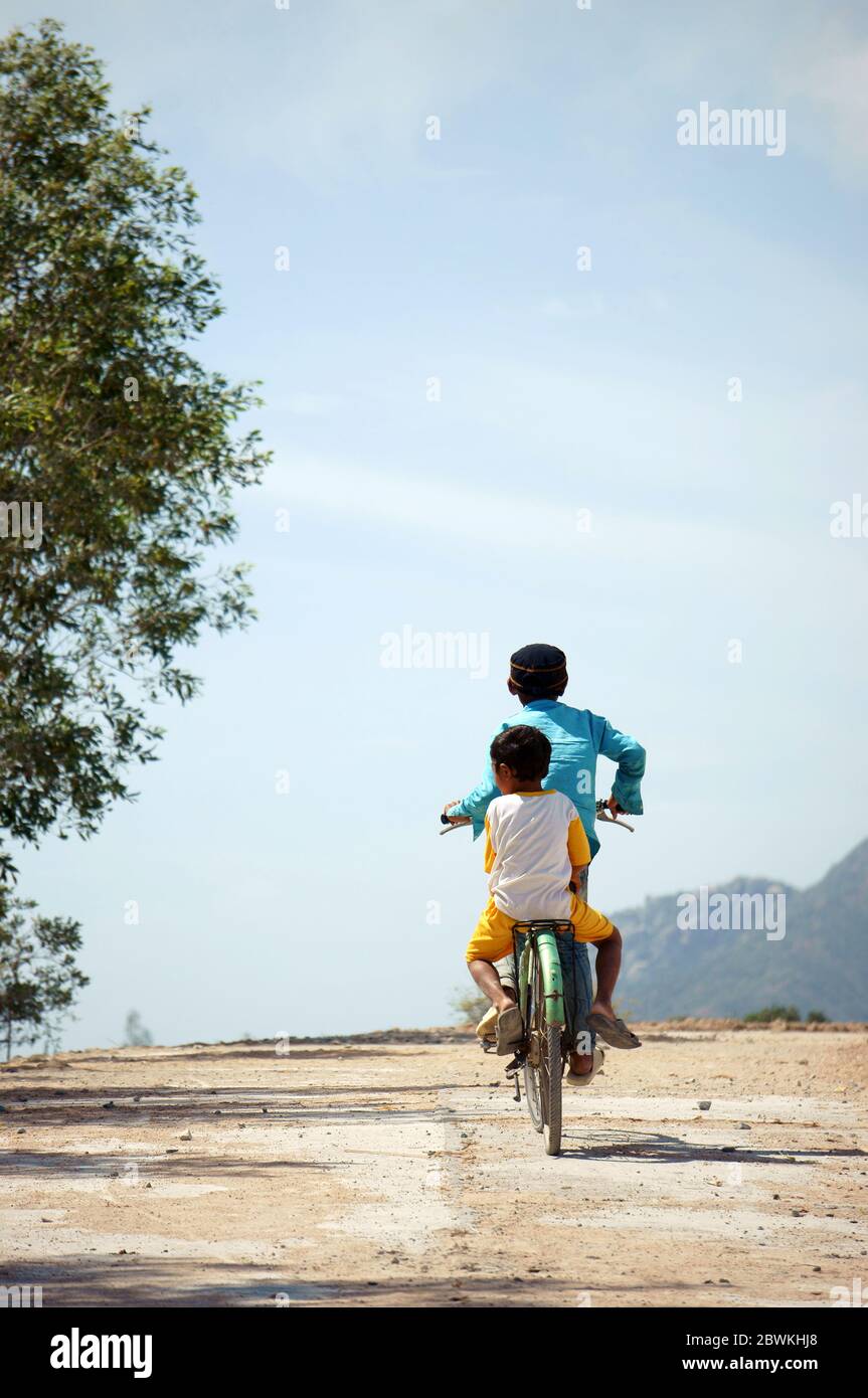 Boy ride a bike go up slope on path of Vietnam deserted countryside, two Vietnamese children enjoy outdoor activity under sun,dirty clothing, wear hat Stock Photo