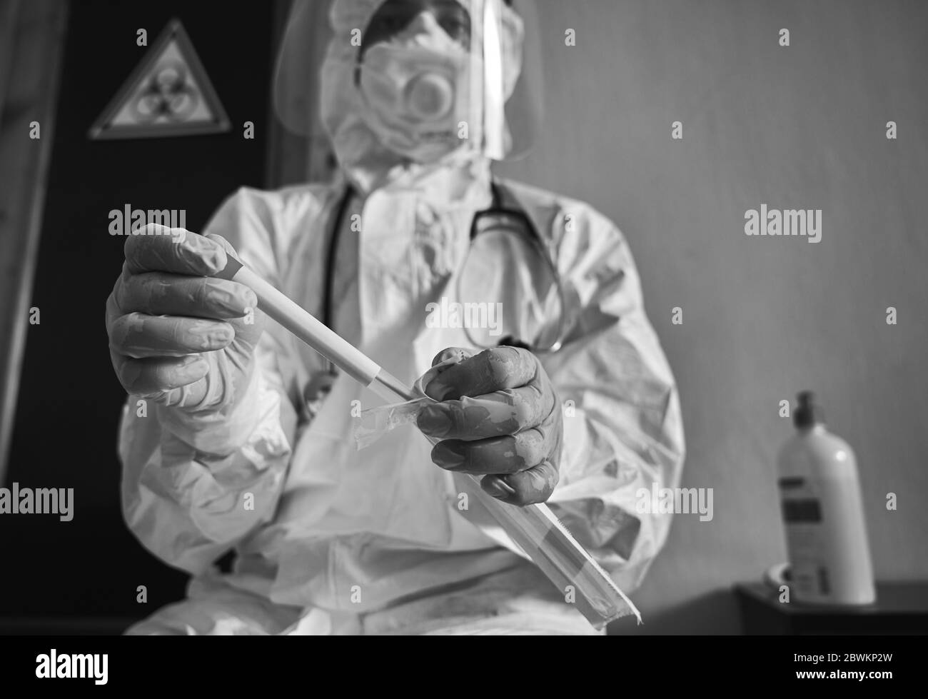 Medical worker holding COVID-19 swab collection kit, wearing white PPE protective suit, face mask, gloves, test tube for taking OP NP patient specimen sample, biohazard sign on background Stock Photo