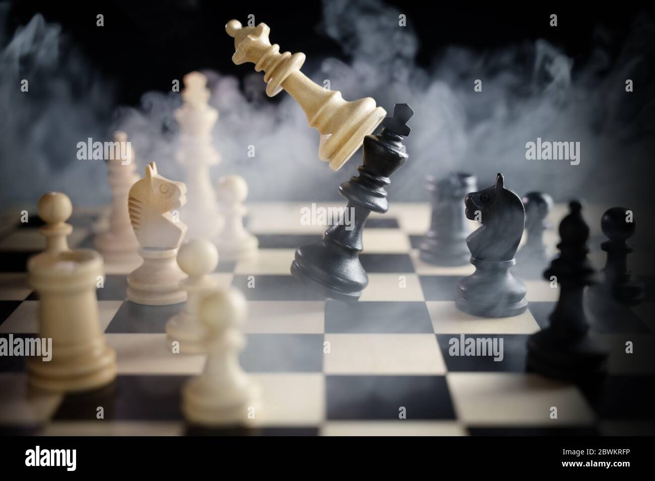 chess queen beats king between other pieces on the chessboard, much smoke over the battle,  against a dark background, concept for aggression, success Stock Photo