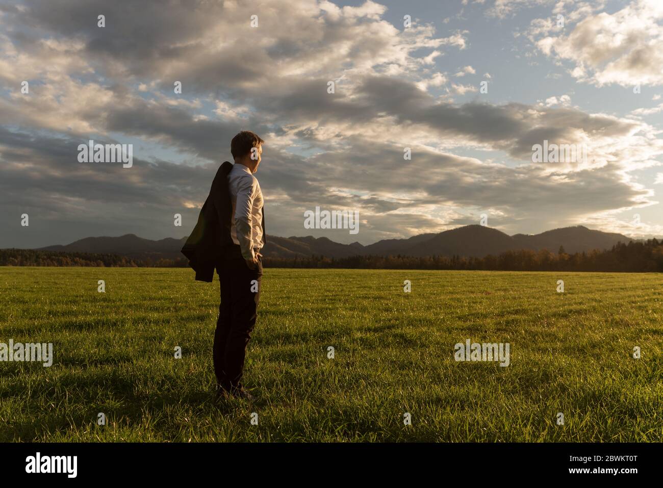 Young businessman with his suit jacket over the shoulder, standing in beautiful green meadow under dramatic evening sky gazing into the distance. Stock Photo