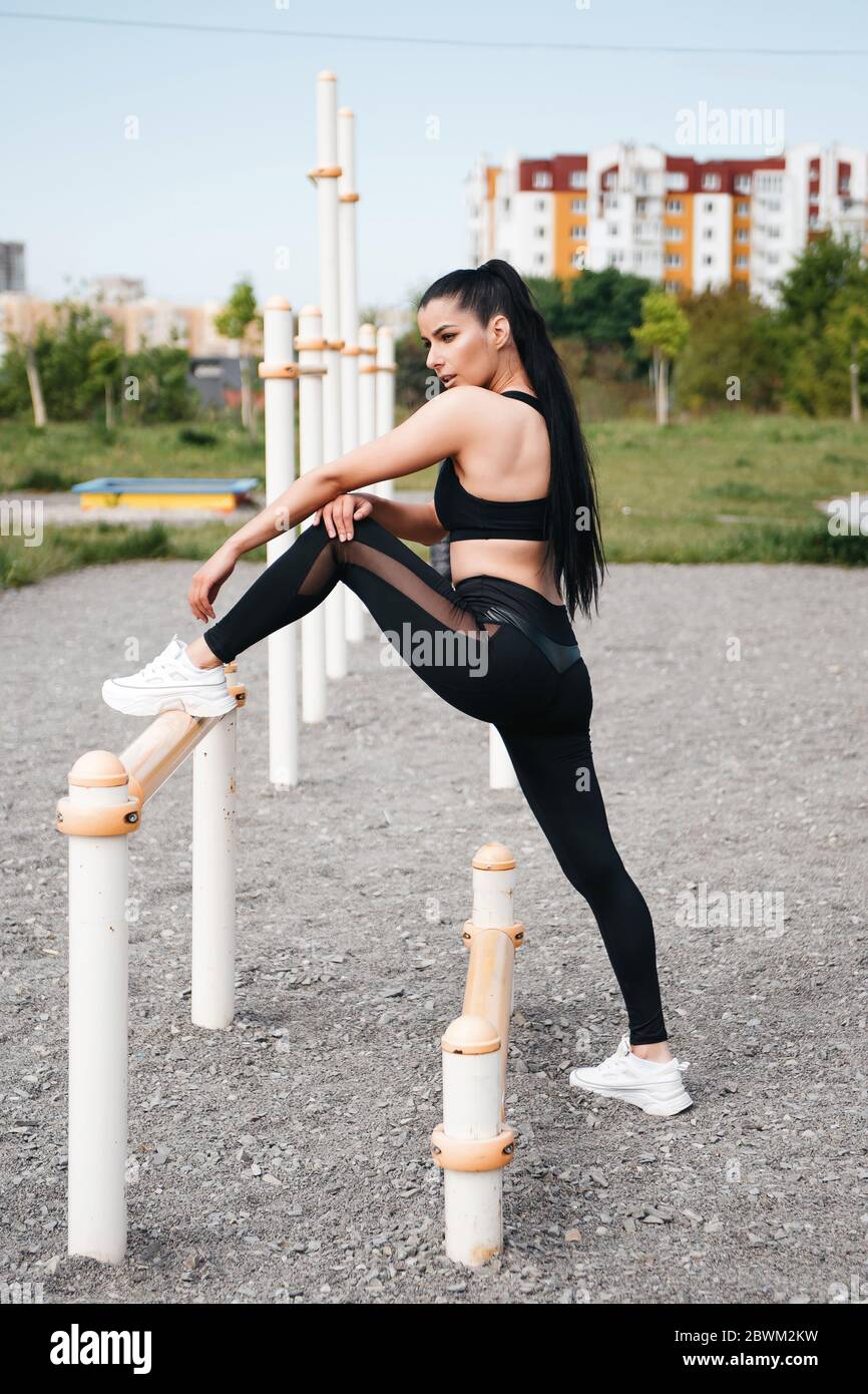 Young brunette in sportswear doing stretching before fitness workout. A woman keeps one leg on a horizontal bar on the sports ground. Sports concept Stock Photo