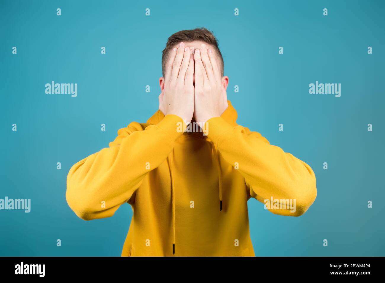 teenager in a sweatshirt on a blue background covers his face with his