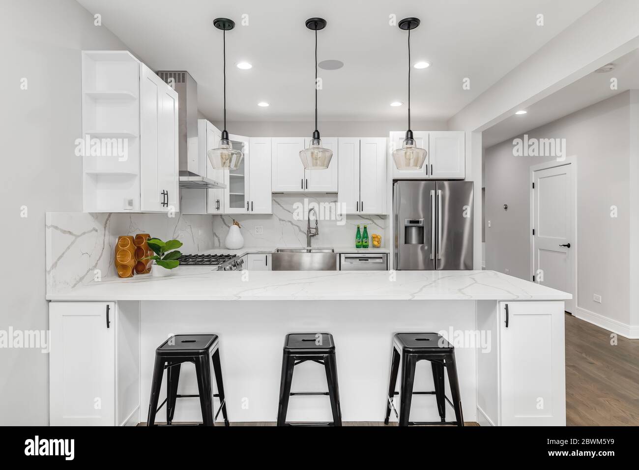 A new, modern all white kitchen with black lights hanging from the ceiling and black bar stools sitting at the counter top for an eating area. Stock Photo