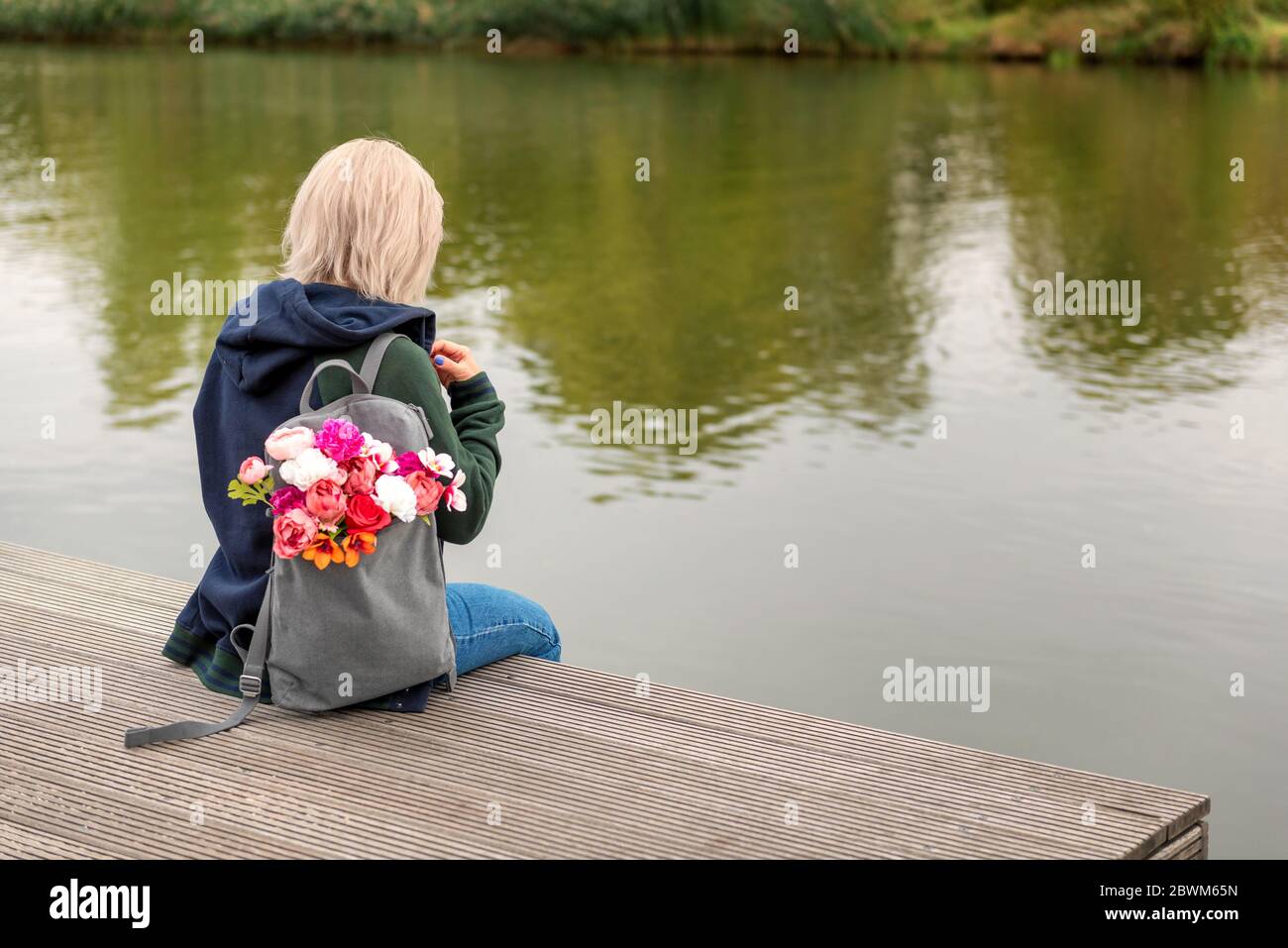 Photo from the back of young blonde dreamy woman with bouquet of flowers in grey backpack near a river. romantic mood Stock Photo