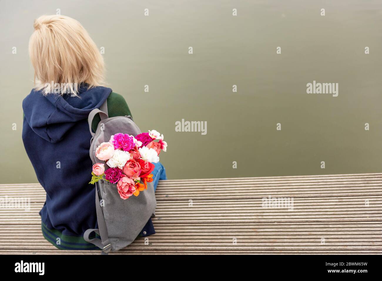 Photo from the back of young blonde dreamy woman with bouquet of flowers in grey backpack near a river. romantic mood Stock Photo
