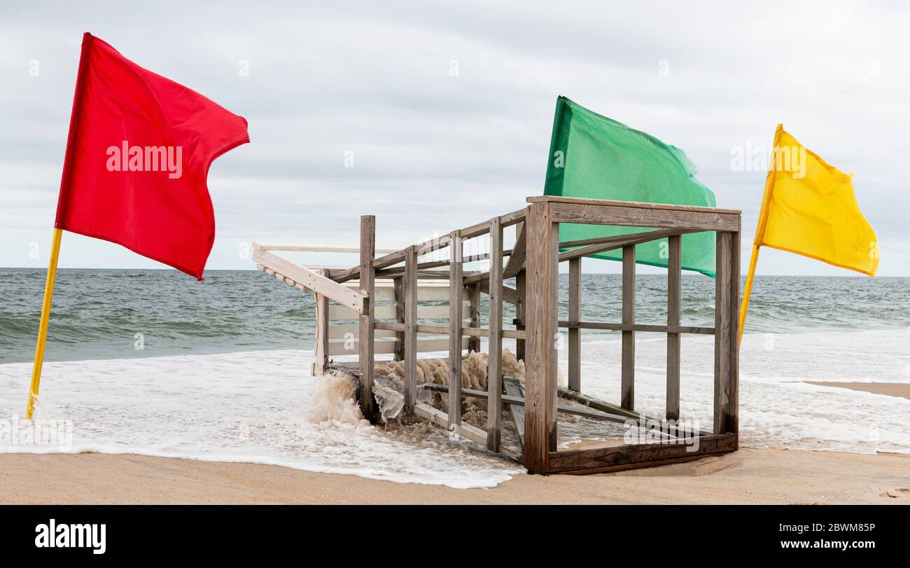 The ocean is overtaking the land under a lifeguar stand which is lying on the sand with red, yellow and green flags up next to it. Stock Photo