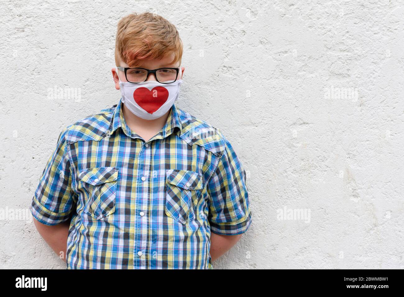 Red-haired boy with glasses and mask with printed red heart. A boy in a plaid shirt and surgical mask stands against a wall background. Stock Photo