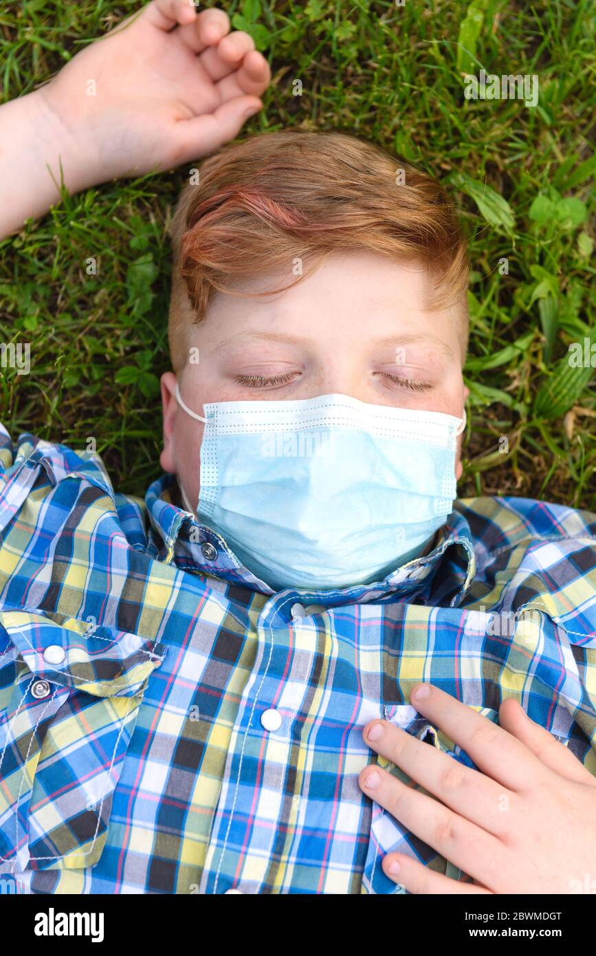 Portrait of a young boy with surgical mask lying on the grass in the park. Close-up of a little boy in a plaid shirt relaxing on the grass. Stock Photo