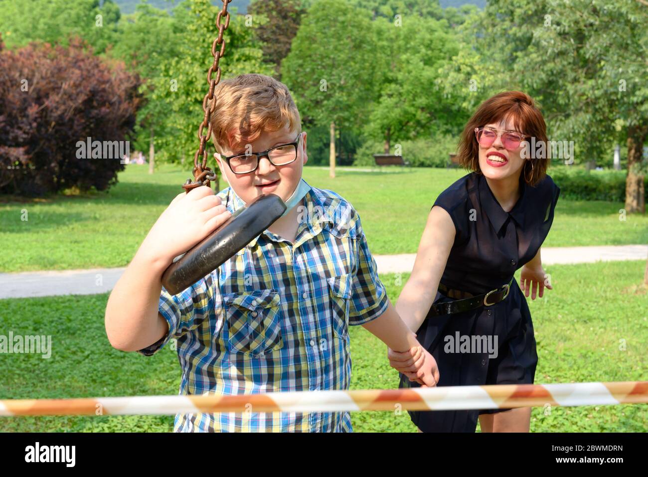 Young boy wearing eyeglasses and surgical mask dressed in a plaid shirt plays with an adult girl in front of a red and white striped barrier tape arou Stock Photo