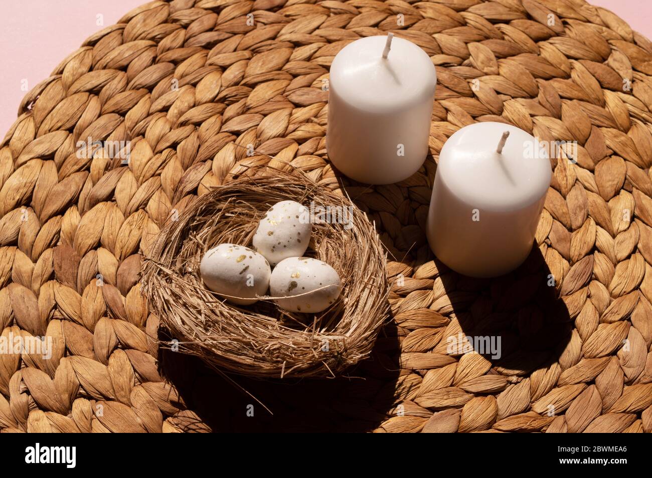 Creative composition of two white candles and three tiny eggs decorated with golden spots inside nest on the light brown mat, handmade from water Stock Photo