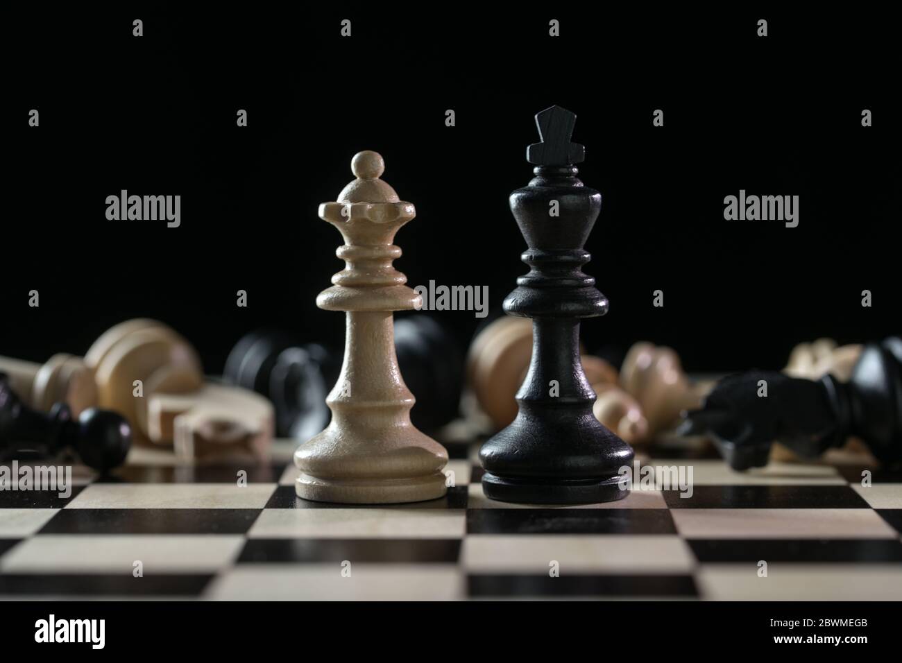 Chess pieces queen and king in front of a of fallen chessmen after a battlefield on a chessboard against a black background, concept of abuse of power Stock Photo