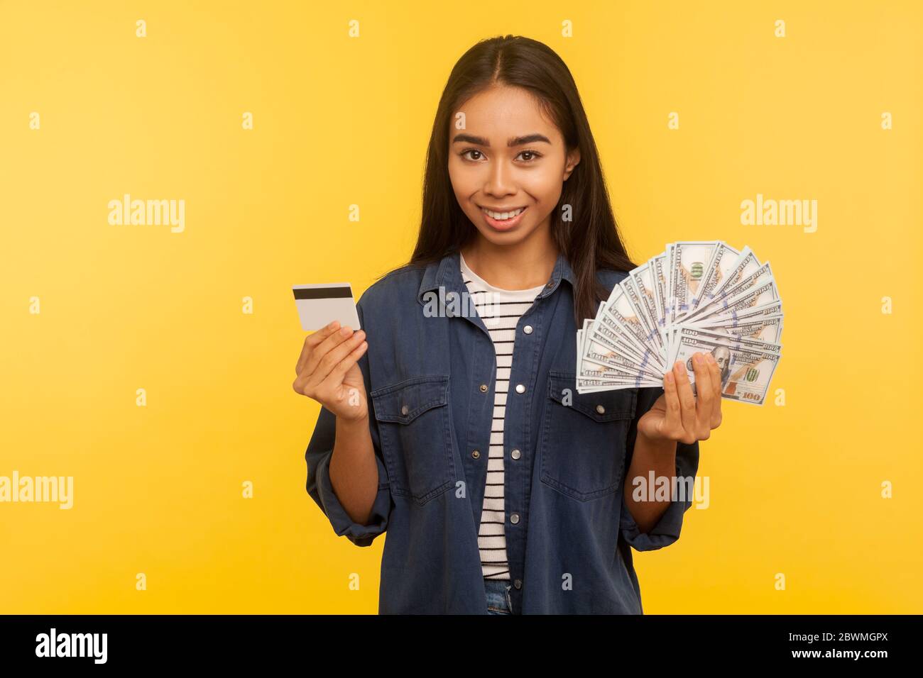 Bank deposit, financial savings. Portrait of happy girl in denim shirt holding credit card and dollar banknotes, looking at camera with toothy smile. Stock Photo