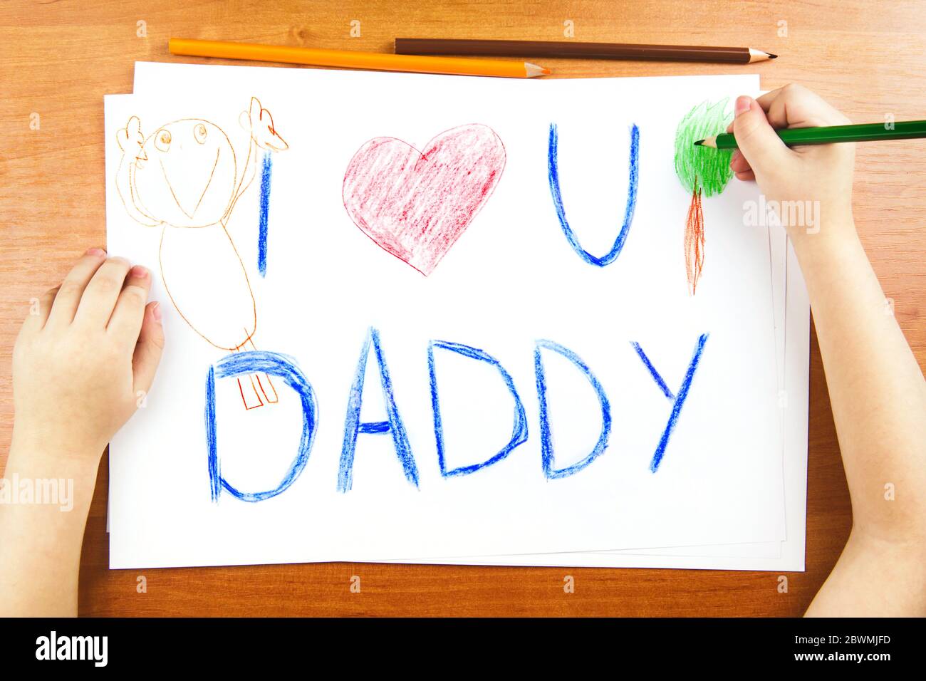 Fathers day concept. Childs hands drawing card with words I Love U Daddy, and red heart, and color pencils on wooden desk. Stock Photo