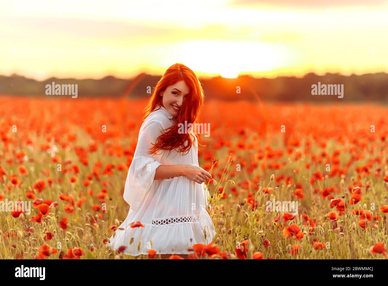 Happy redhead smiling woman in white dress on field of poppies at warm ...