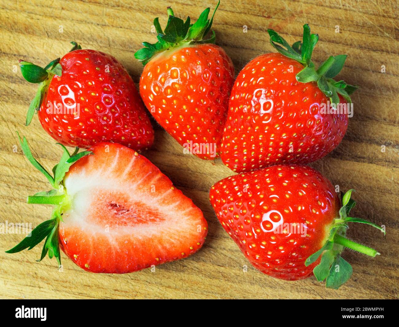 Four whole  strawberries and half a strawberry on a wooden chopping board Stock Photo