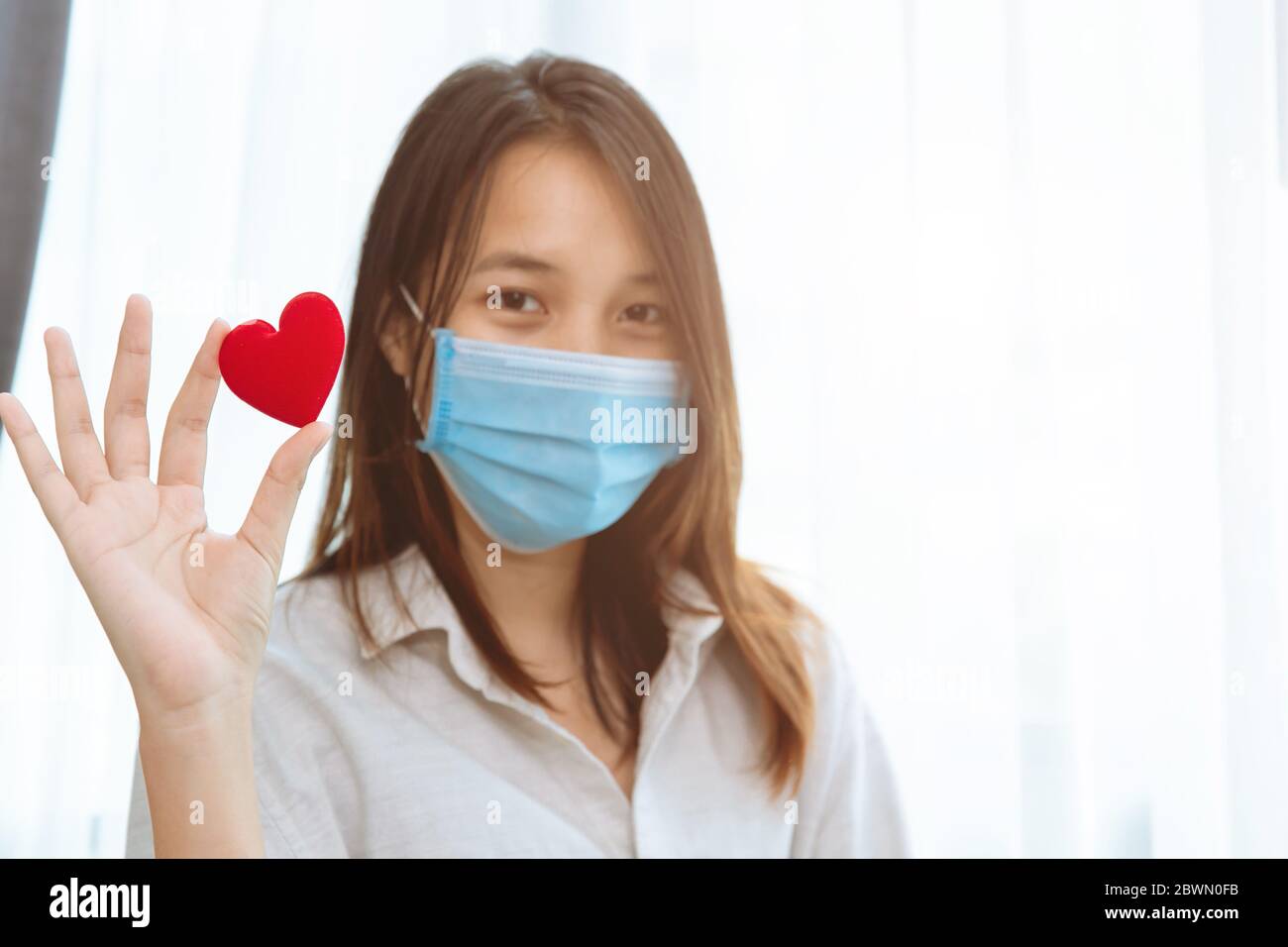 Cute Asian girl teen wear face mask hand holding red heart for love care together stay healthy and protect Coronavirus(Covid-19) spreading concept. Stock Photo