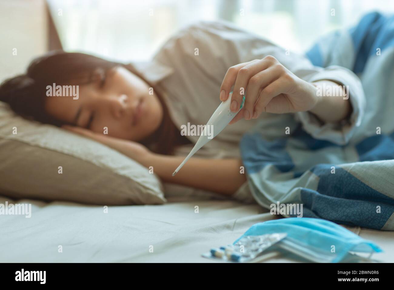 Asian teen infected with Covid-19 flu sick lying in bed due to a Corona virus pandemic, anxiously measuring check body temperature with digital thermo Stock Photo